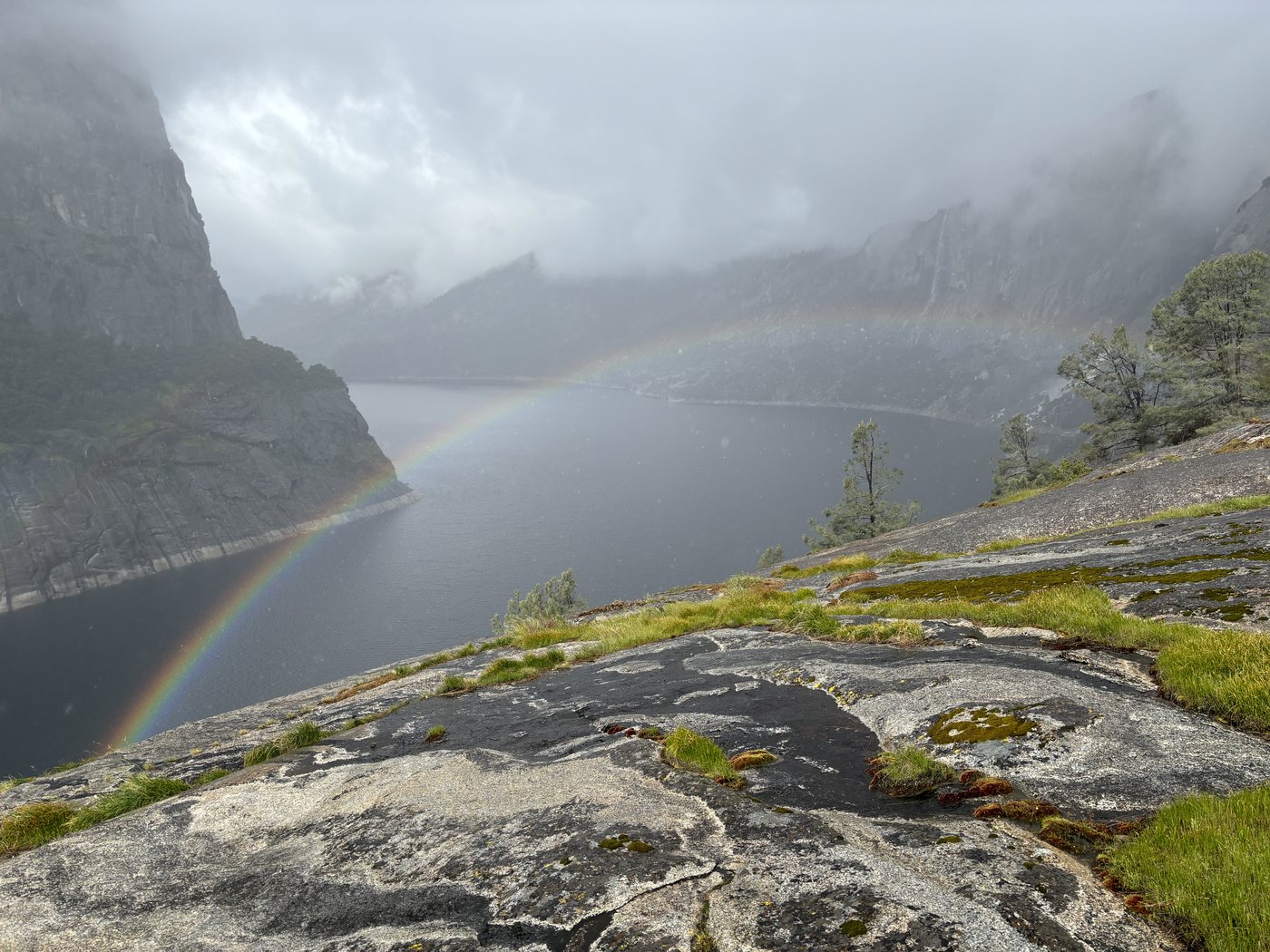 A full rainbow arcing across Hetch Hetchy Reservoir in the rain, low cloud sitting on the granite walls on both sides