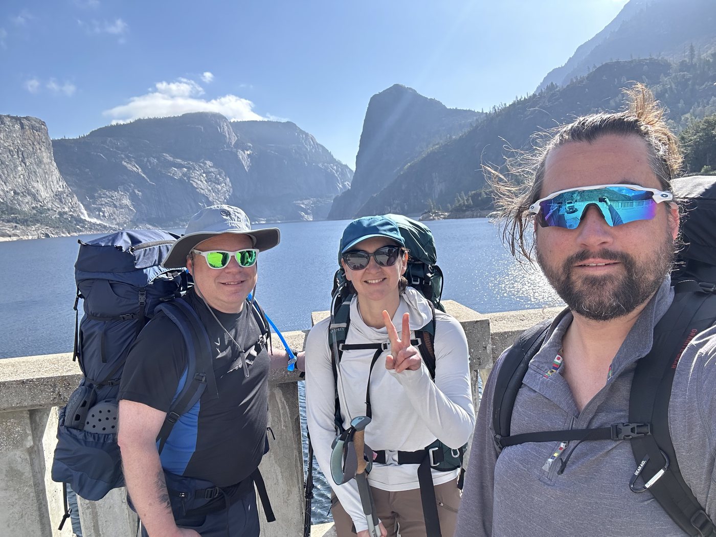 Three backpackers with full packs standing on top of O'Shaughnessy Dam, the reservoir and granite walls behind them under a bright blue sky
