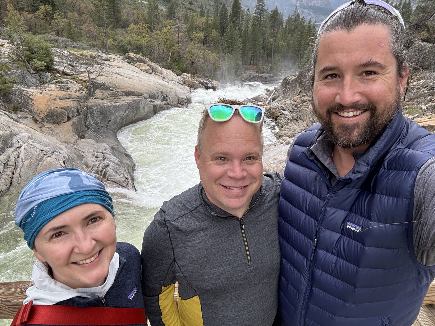 Three hikers smiling closely together for a selfie on rock beside a rushing river, spray rising behind them