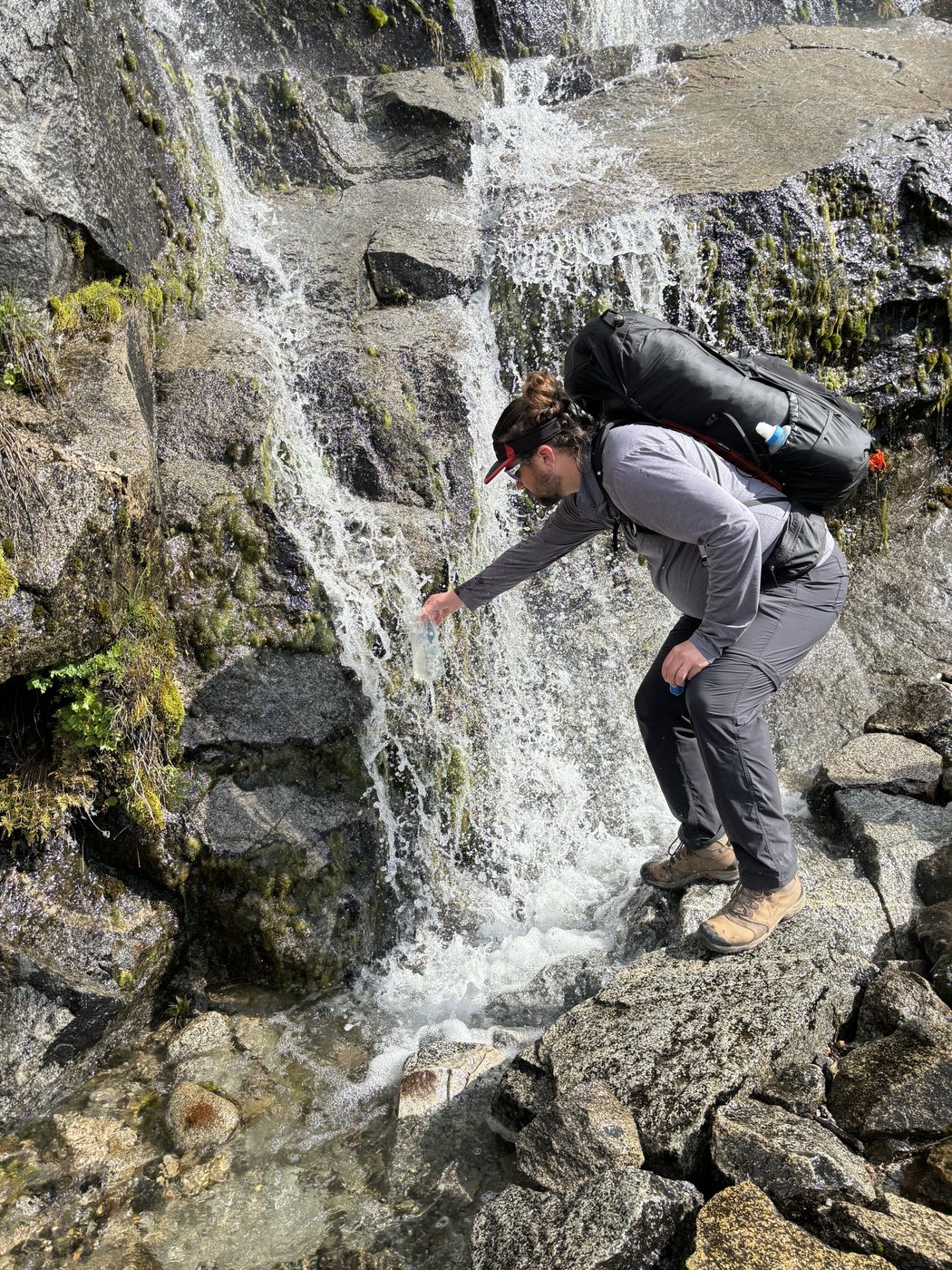 A hiker in a grey shirt stepping across wet rock beneath a small waterfall that falls directly across the trail