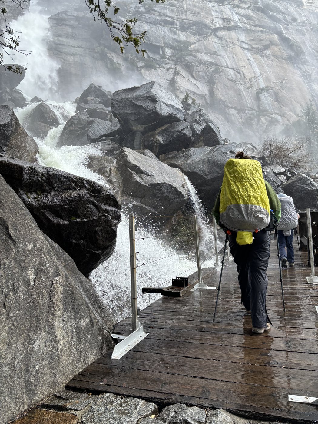 A backpacker with a yellow pack cover crossing a wet wooden footbridge while whitewater surges through the cable railing beside them, spray and mist filling the air