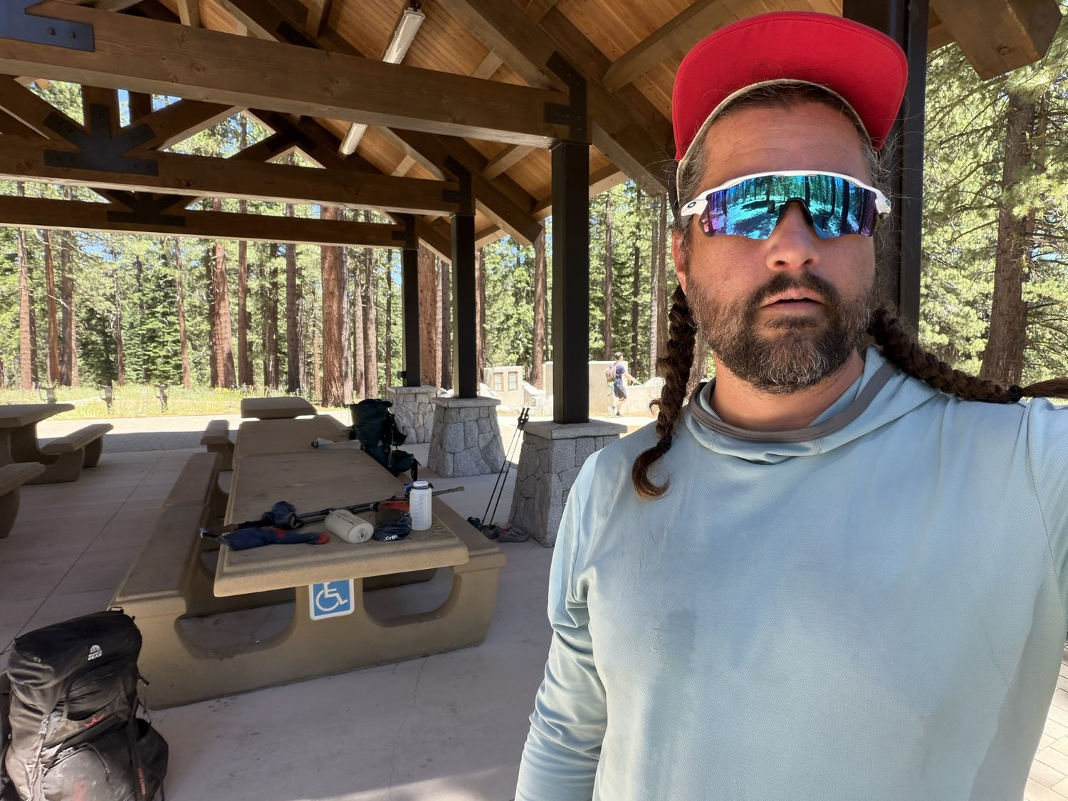 A hiker at a shaded trailhead picnic shelter with backpacks and gear on the table behind him