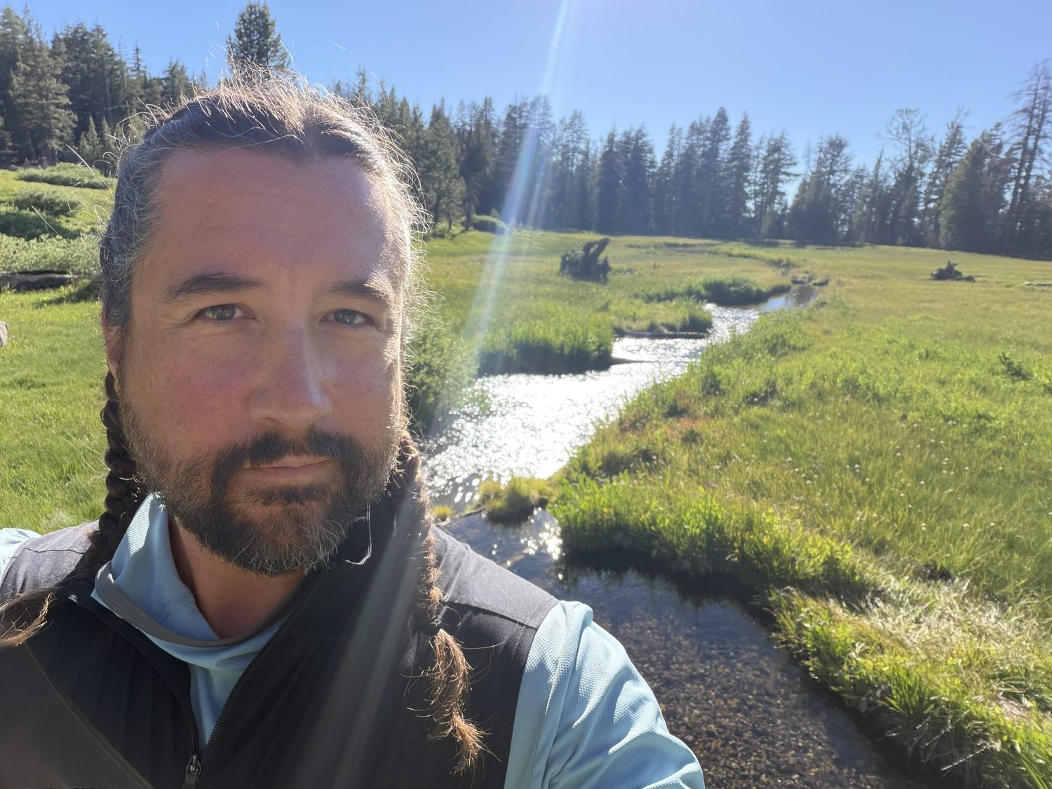 A hiker's selfie with a green meadow and a winding inlet stream leading toward Gray Lake in the background