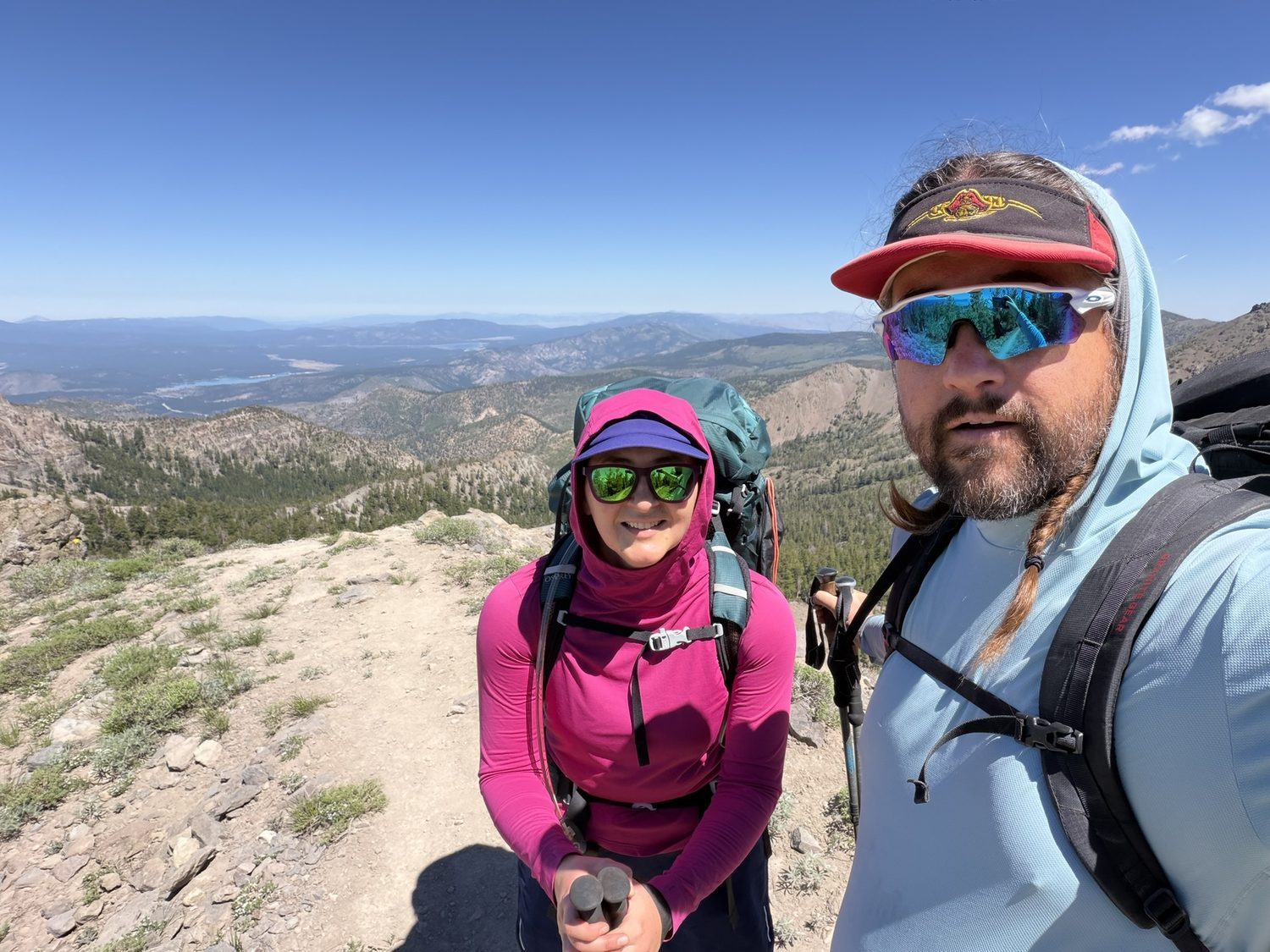 Two hikers at a high, barren summit with mountains and haze stretching to the horizon