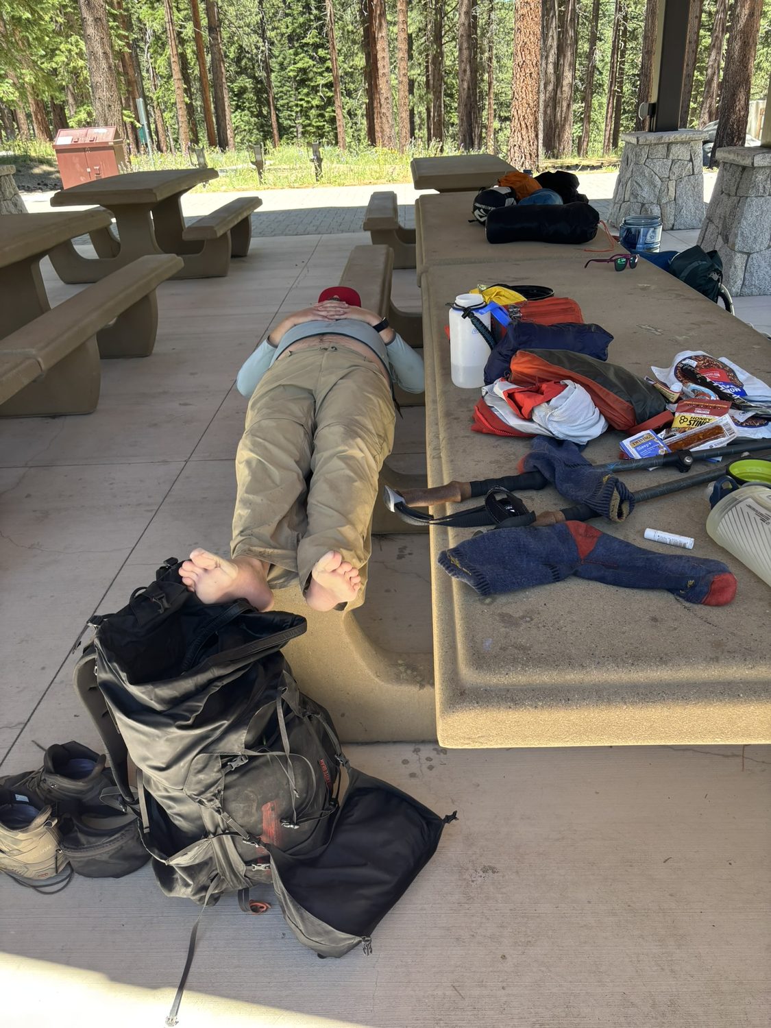 A barefoot hiker lying flat on a bench with feet up and gear drying on the picnic table at the Spooner trailhead shelter