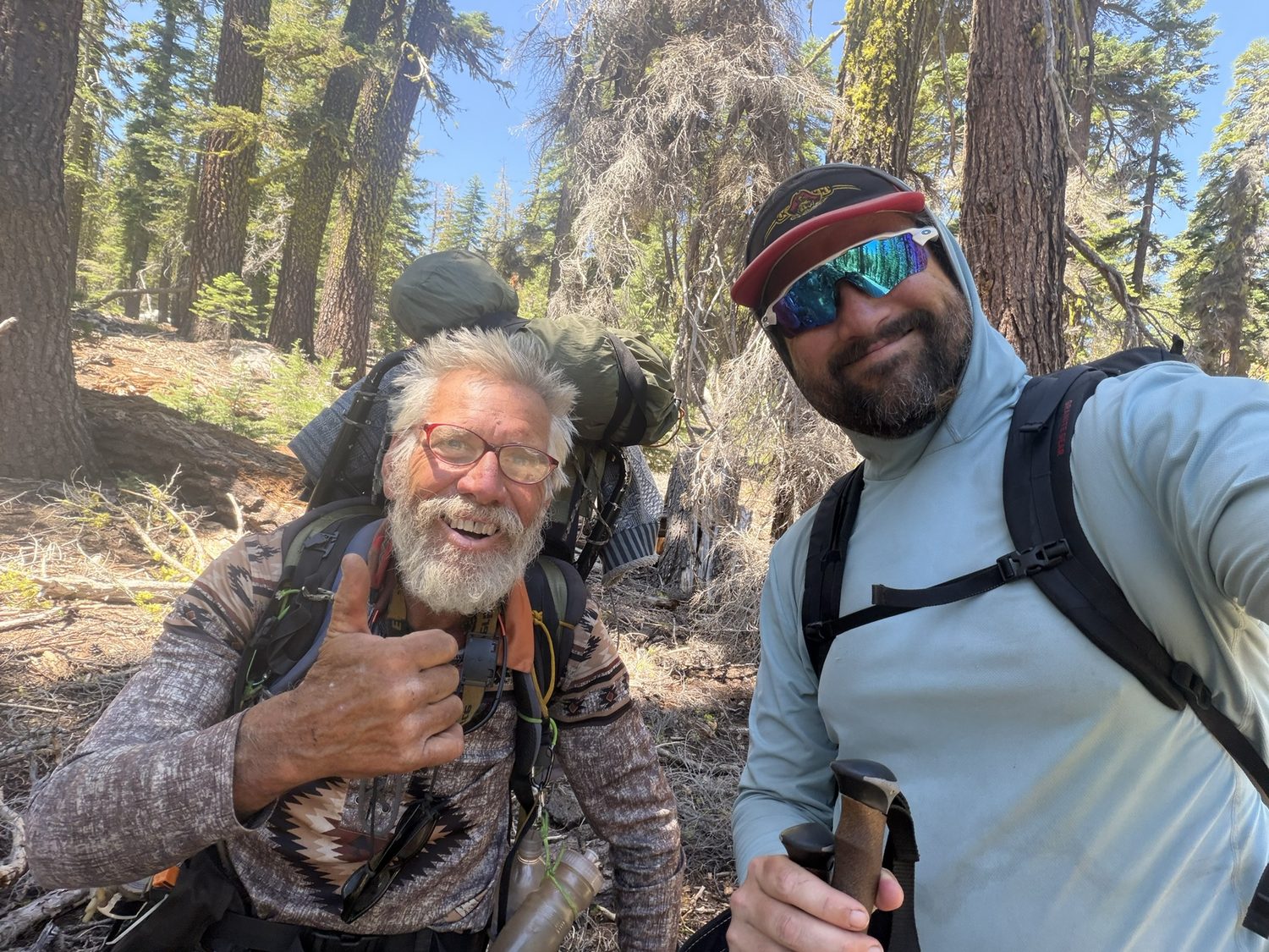 Two hikers taking a selfie in the forest, one with a gray beard and a large backpack giving a thumbs-up