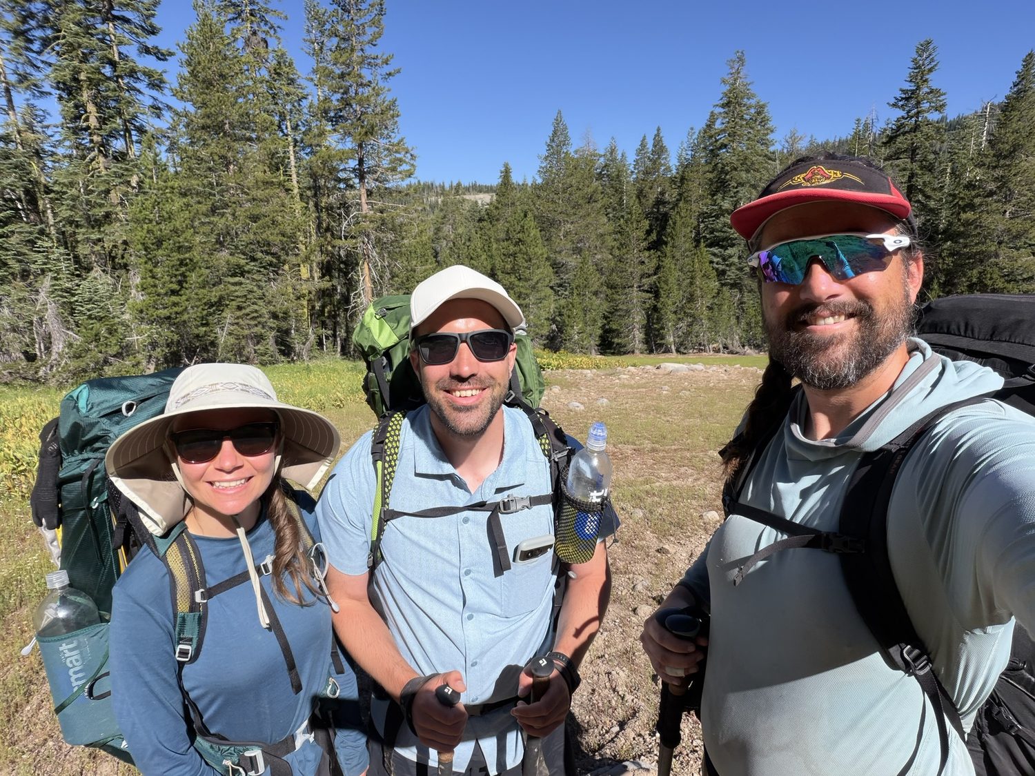 Three hikers with full backpacks smiling together on the trail in a green meadow