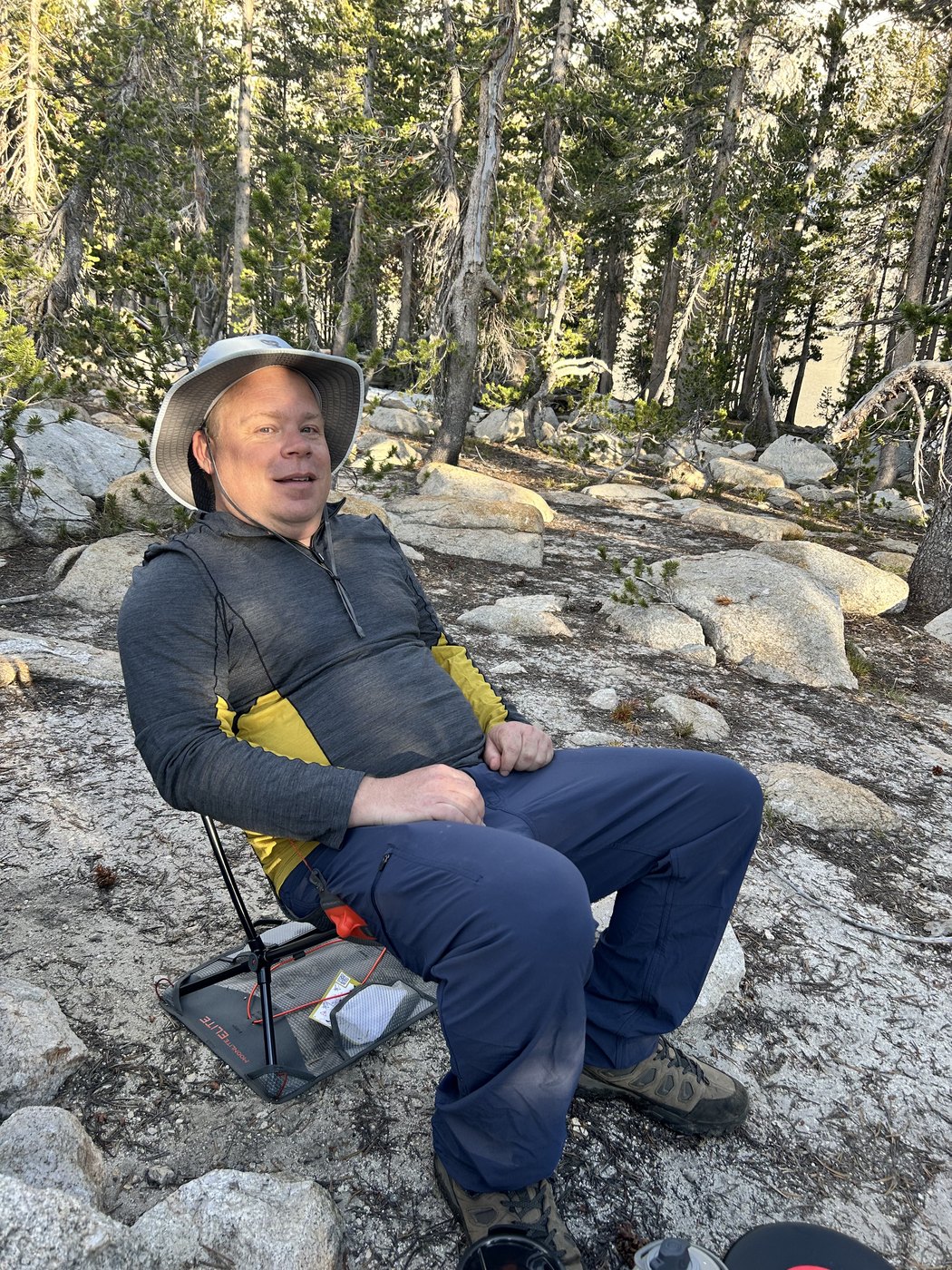 A hiker in a wide-brimmed sun hat sitting back in a low folding camp chair on a granite slab among pines, entirely at ease in the late sun