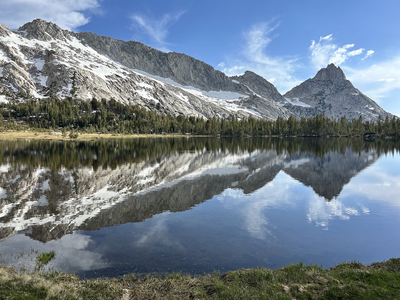 A high alpine lake gone perfectly still in evening light, doubling a snow-streaked granite ridge, a sharp peak, and a sky of long white clouds in a flawless reflection