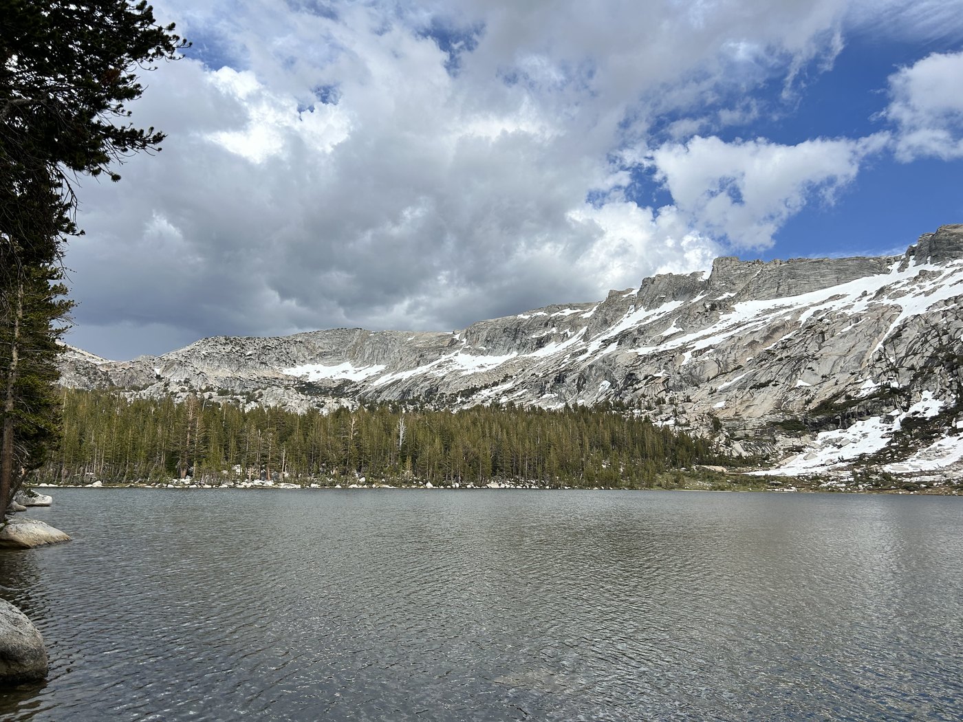An alpine lake in the late afternoon with a long granite ridge across the water carrying broad unbroken fields of old snow above a dark band of trees