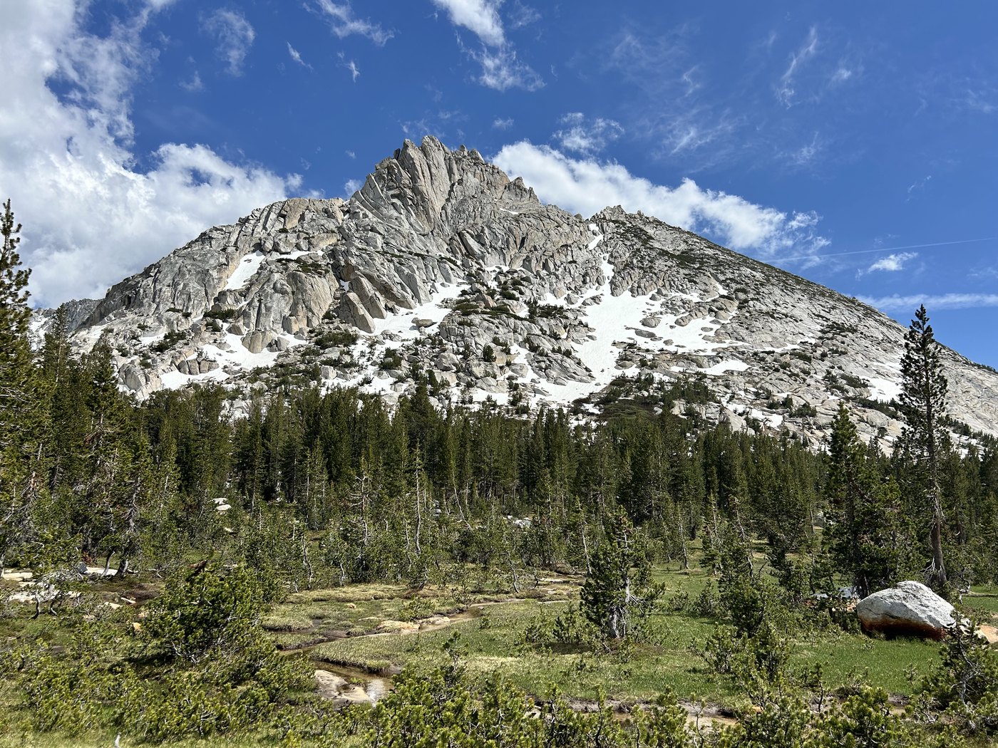 A steep grey granite peak with patches of snow on the flanks standing above a foreground of low scrub and scattered pines under a blue sky with high cloud