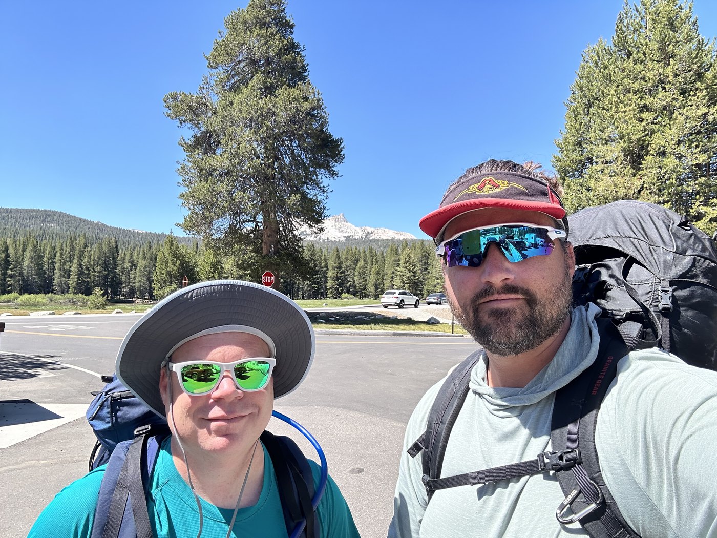 Two backpackers taking a selfie in a paved trailhead parking area, one in a red cap and one in a wide-brimmed sun hat, both wearing mirrored sunglasses with packs on and pines behind them