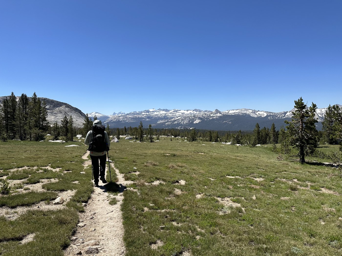 A backpacker walking away along a sandy trail through open alpine meadow, with a granite dome and a long line of distant snowy peaks across the horizon